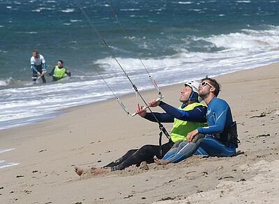 Kiteschüler mit Kitelehrer sitzen am Strand und üben die Kitesteuerung