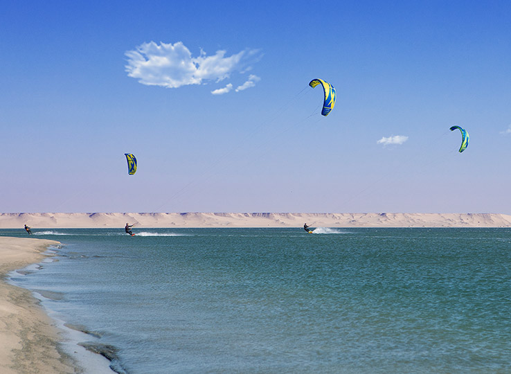 Speedspot kitesurf sulla spiaggia turchese con tre kitesurfisti e aquiloni colorati contro paesaggio desertico