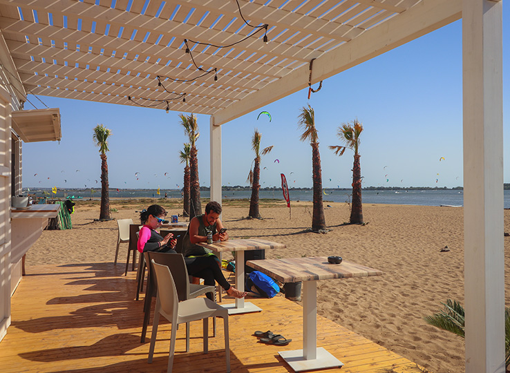 Café sulla spiaggia Sizilien Kitecenter con ospiti sotto pergolato, palme e spiaggia kitesurf sullo sfondo