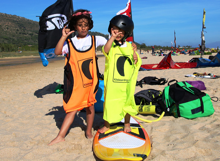 Two children in kiteboarding vests on beach with kite equipment and flags - kids kitesurfing lessons at beach destination